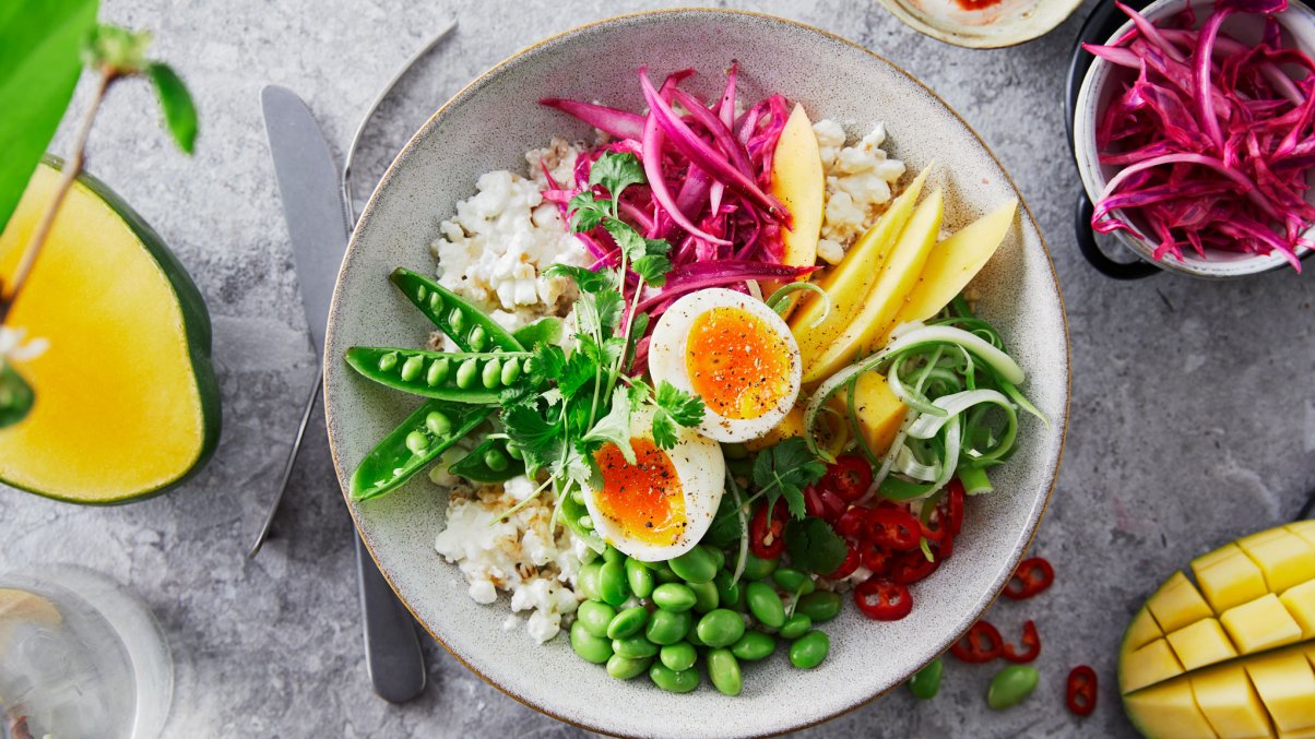 Bildet er tatt rett ovenfra ned i en rund, stor skål. I skålen ligger det cottage cheese i bunn med masse friske farger rundt. Det er oppskjært gul, mango. Rød chili delt i skiver, syltet rosa rødløk, grønne sukkererter delt på langs, grønne bønner og grønn vårløk.